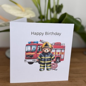 May include: A white birthday card with the words "Happy Birthday" features a cartoon illustration of a teddy bear dressed as a firefighter in front of a red fire engine. The bear wears a yellow helmet and a black and yellow uniform.