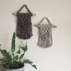 May include: Two macrame wall hangings with wooden dowels. The one on the left is brown and the one on the right is beige.