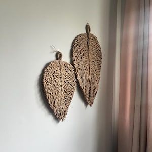 Op de afbeelding: Twee geweven wanddecoraties in de vorm van bladeren, gemaakt van natuurlijke vezels. Het grotere blad is bruin en het kleinere blad is lichtbruin. De bladeren hangen aan een witte muur.