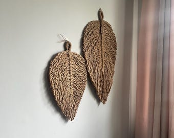 May include: Two woven leaf wall hangings made of natural fibers. The larger leaf is brown and the smaller leaf is a lighter brown. The leaves are hanging on a white wall.