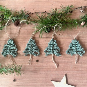 May include: Four green macrame Christmas tree ornaments hanging from twine on a wooden surface. The ornaments are handmade and have a rustic, natural look.