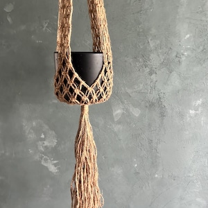 Puede incluir: Un colgador de plantas hecho de yute tejido de color beige. El colgador presenta una maceta negra suspendida dentro de una red de macramé, con largas borlas con flecos en la parte inferior. El fondo es una pared gris texturizada.
