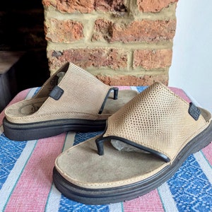 Può includere: Un paio di sandali in pelle color cuoio con un design a rete e una suola spessa nera. I sandali sono su una superficie di tessuto a righe.