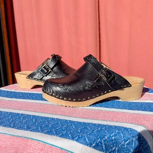 Könnte beinhalten: Ein Paar schwarze Ledersandalen mit Holzsohle und Schnallenriemen. Die Sandalen haben ein florales Design, das in das Leder geschnitzt ist.