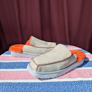 Puede incluir: Un par de zapatillas de ante gris con forro de piel naranja. Las zapatillas tienen una suela azul claro y costuras rojas alrededor de los bordes.