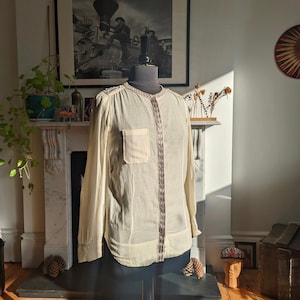 May include: A cream-colored, long-sleeved blouse with a button-up front and a small pocket on the left chest. The blouse has a beaded trim down the front and around the collar.