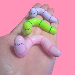 May include: Three colorful, smiling worm figurines in shades of pink, green, and purple are held in a hand against a pink background. The worms have painted details, including stripes and happy faces, adding a whimsical touch.
