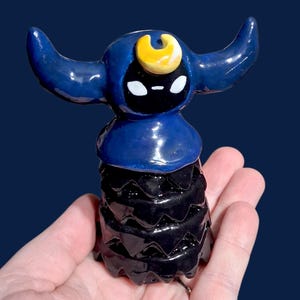May include: A handmade ceramic figurine with a dark blue hood and a yellow crescent moon detail. The figurine has a black face with white eyes and a black, textured body. The figurine is held in a hand against a dark blue background.