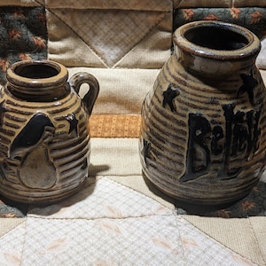 May include: Two tan, ribbed ceramic vessels with dark brown glaze accents. The smaller vessel features a handle and a pear-shaped relief. The larger vessel displays the word "BELIEVE" in dark lettering. Both items are decorated with small star motifs.