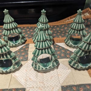 May include: Six ceramic Christmas tree-shaped candle holders. The candle holders are green with white detailing and a star on top. The candle holders are arranged on a patterned placemat.