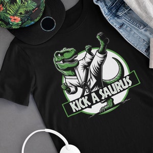 May include: Black t-shirt with a green cartoon dinosaur wearing a white karate uniform and kicking. The dinosaur is in a circle with the text "Kick a Saurus" in green letters.