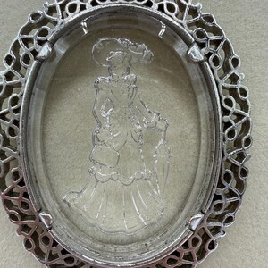 May include: An oval, silver-toned pendant with a clear, three-dimensional cameo of a woman in a dress and hat. The pendant has an ornate, openwork border. The cameo depicts a woman holding a parasol.