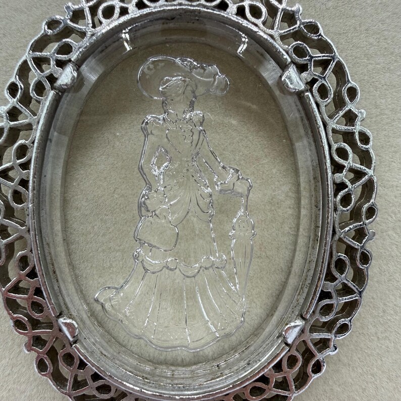 May include: An oval, silver-toned pendant with a clear, three-dimensional cameo of a woman in a dress and hat. The pendant has an ornate, openwork border. The cameo depicts a woman holding a parasol.