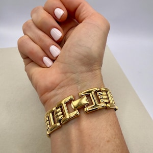 May include: A gold-toned bracelet with a rectangular clasp and geometric links. The bracelet is worn on a wrist, showcasing its polished finish and intricate design. The bracelet's links are approximately 0.75 inches wide.