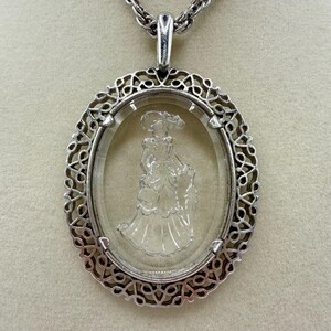 May include: Silver-toned oval pendant necklace. The pendant features a clear, oval-shaped glass with an etched image of a woman in a dress and hat. The pendant is framed by a decorative, openwork silver border and hangs from a silver chain.