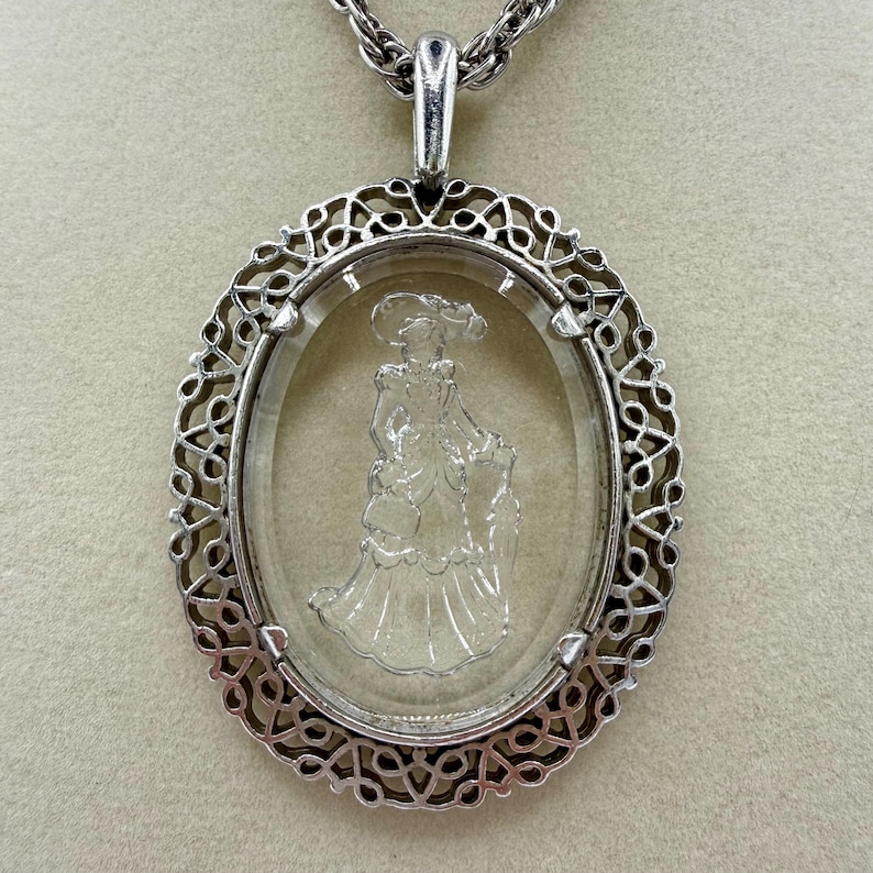 May include: Silver-toned oval pendant necklace. The pendant features a clear, oval-shaped glass with an etched image of a woman in a dress and hat. The pendant is framed by a decorative, openwork silver border and hangs from a silver chain.