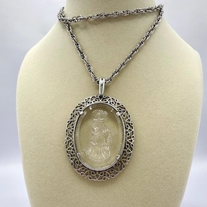 May include: Silver-toned necklace with an oval pendant. The pendant features a clear, etched design of a woman in a dress, framed by a decorative silver border. The chain is a linked design, and the necklace is displayed on a mannequin.