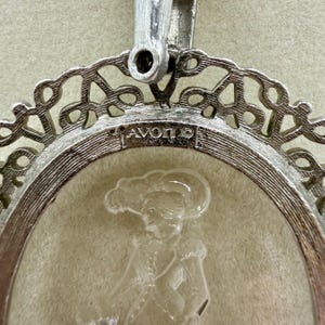 May include: Silver-toned pendant with an oval, clear center featuring a cameo of a person in a hat. The pendant has a decorative filigree border and the word "AVON" is visible. The clasp is at the top.