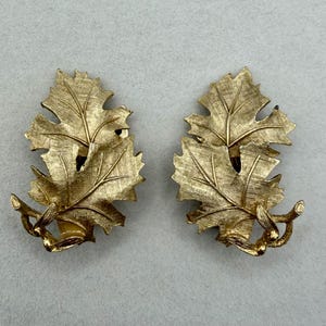 May include: A pair of gold-toned leaf-shaped clip-on earrings. Each earring features three textured leaves with detailed veining, attached to a small branch with a cluster of berries. The earrings have a vintage aesthetic.