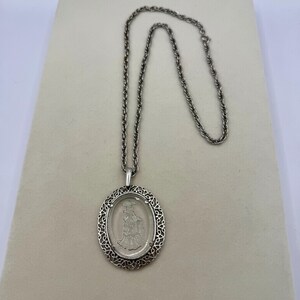 May include: Silver-toned pendant necklace featuring an oval pendant with a detailed, etched design of a figure. The pendant is encased in a decorative, openwork frame and hangs from a matching chain. The necklace is displayed on a neutral background.