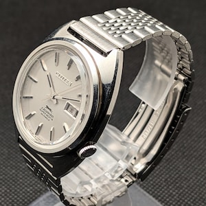 May include: A silver-toned Citizen Leopard Super Beat 28 Jewels wristwatch. The watch features a silver dial with silver hands and markers, a date window, and a stainless steel mesh band. The watch is displayed on a clear stand.