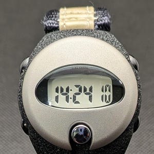 May include: A digital watch with a black strap and a silver-coloured face. The watch displays the time as 14:24 and the date as 10. The watch band is black and the watch face has the text "ALBA SPOON".