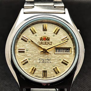 以下が含まれることがあります： 金色の文字盤が付いたシルバーカラーのオリエントの腕時計。文字盤には曜日と日付が表示され、金色の針とマーカーが付いています。時計バンドはシルバーカラーの金属製です。文字盤には「Orient」、「Crystal」、「21 Jewels」と書かれています。