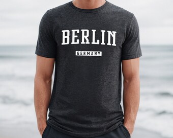 May include: A dark gray t-shirt with the word "BERLIN" in white block letters and the word "GERMANY" in a white rectangle below. The shirt is being worn by a person standing in front of a beach.