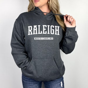 Peut inclure: Un sweat &agrave; capuche gris fonc&eacute; avec le texte "RALEIGH NORTH CAROLINA" en blanc.