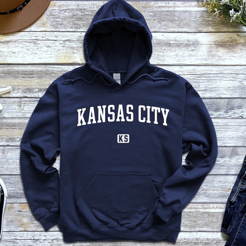 Pu&ograve; includere: Una felpa con cappuccio blu navy con il testo "KANSAS CITY KS" in bianco.