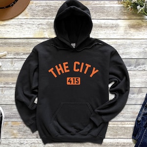 Op de afbeelding: Zwart hoodie met een oranje afbeelding met de tekst "THE CITY 415".