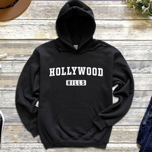 May include: Black hooded sweatshirt with white text that reads "HOLLYWOOD HILLS" in a collegiate font.