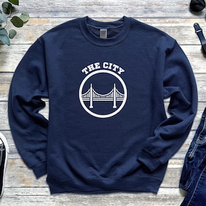 Op de afbeelding: Een marineblauw sweatshirt met een witte afbeelding van de Golden Gate Bridge en de tekst "THE CITY".