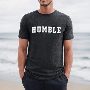 May include: Dark gray t-shirt with the word "HUMBLE" printed in bold white letters. The shirt is a crew neck style and appears to be made of a soft material. The background is a blurred image of a beach and ocean.