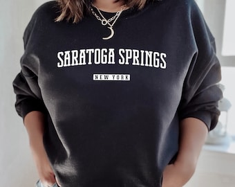 Puede incluir: Sudadera negra con texto blanco que dice "SARATOGA SPRINGS NEW YORK".