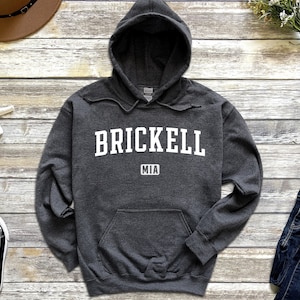 Puede incluir: Sudadera con capucha gris oscuro con la palabra "BRICKELL" en letras blancas en el pecho, con "MIA" debajo. La sudadera tiene un bolsillo delantero y una capucha con cordón. La prenda está hecha de un material suave.