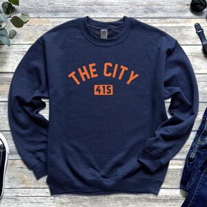 Op de afbeelding: Marineblauwe sweatshirt met de tekst "THE CITY" in oranje gebogen letters boven het nummer "415" in een rechthoekige oranje doos. De sweatshirt heeft een ronde hals.