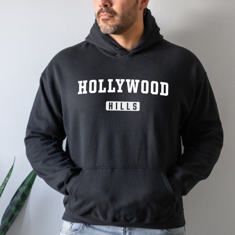 May include: Black hooded sweatshirt with white text that reads "HOLLYWOOD HILLS" in a vintage collegiate style font.