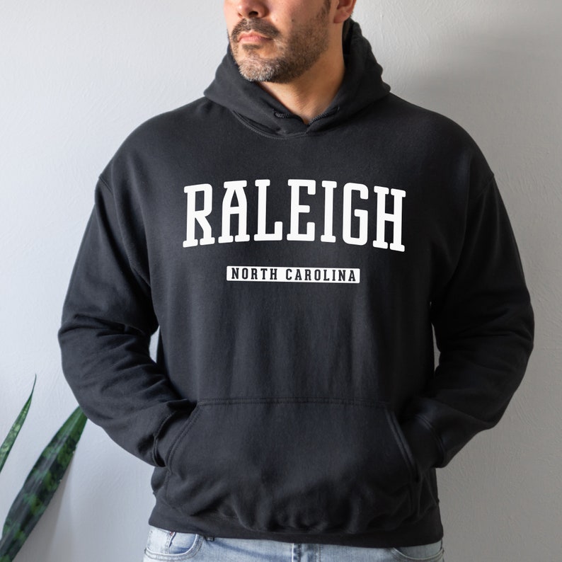 Peut inclure: Sweat-shirt &agrave; capuche noir avec un texte blanc qui dit "RALEIGH NORTH CAROLINA".