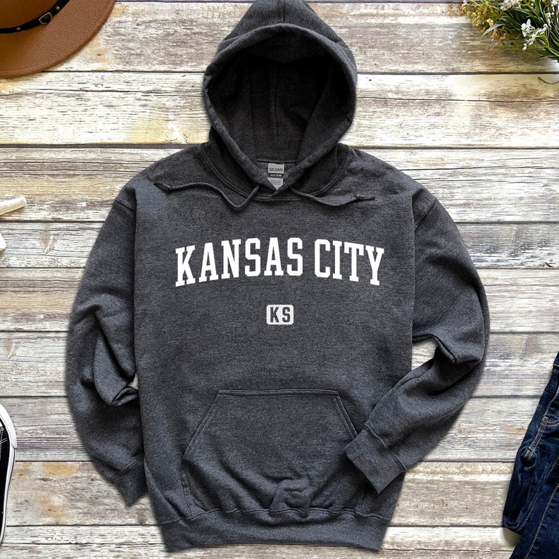 Pu&ograve; includere: Una felpa con cappuccio grigio scuro con il testo "KANSAS CITY KS" in bianco.