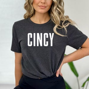 Puede incluir: Una camiseta gris oscuro con la palabra "CINCY" impresa en blanco en la parte delantera.