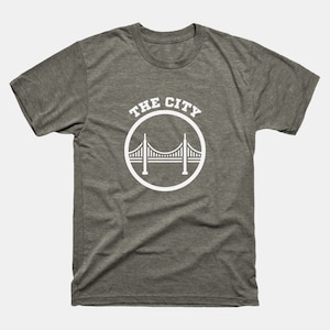 Op de afbeelding: Een heidekleurig T-shirt met een witte cirkelvormige afbeelding met een brug en de tekst "THE CITY".
