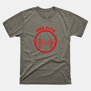 Op de afbeelding: Een heidekleurig t-shirt met een rode cirkelvormige afbeelding van de Golden Gate Bridge en de tekst "THE CITY".