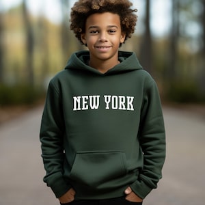以下が含まれることがあります： 前面に白い文字で「NEW YORK」とプリントされた濃い緑色のフード付きスウェットシャツです。