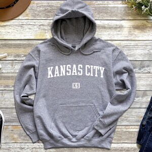 Pu&ograve; includere: Una felpa con cappuccio grigia con la scritta "KANSAS CITY KS" stampata in bianco sul davanti.