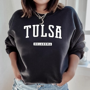 Könnte beinhalten: Schwarzes Sweatshirt mit dem Wort "TULSA" in großen weißen Blockbuchstaben über dem Wort "OKLAHOMA" in kleinerer weißer Schrift. Das Sweatshirt hat einen Rundhalsausschnitt.