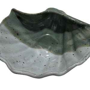 May include: A ceramic dish shaped like a seashell with a scalloped edge. The surface features a two-tone glaze with a glossy white finish speckled with dark brown dots on the lower half and a muted sage green glaze on the upper interior section.