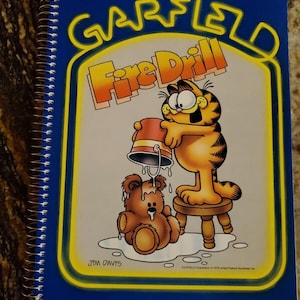 May include: A blue spiral notebook with a yellow border. The cover features a cartoon image of Garfield holding a bucket of water over a teddy bear. The text "GARFIELD Fire Drill" is written in yellow letters on the cover. The artist's name "JIM DAVIS" is written in the bottom left corner.