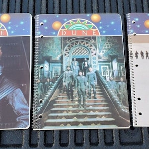 May include: Three spiral bound notebooks with a "Dune" theme. The covers feature images from the 1984 film adaptation of Frank Herbert's novel. The first notebook shows a close-up of Kyle MacLachlan as Paul Atreides. The second notebook shows a group of characters walking up a staircase. The third notebook shows Sting as Feyd-Rautha.