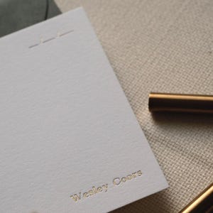 Puede incluir: Una tarjeta de papeler&iacute;a rectangular blanca con el nombre Wesley Coors grabado en letras de papel de oro. La tarjeta incluye un detalle de l&iacute;nea horizontal sutil en la parte superior y se coloca junto a un bol&iacute;grafo dorado met&aacute;lico sobre una tela.